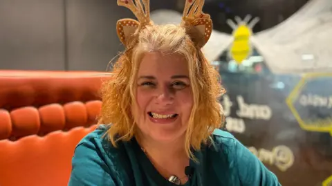 Suzanne Hemmings is wearing a green top and has red hair. She is wearing a reindeer on top of her head.