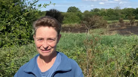 Henry Godfrey-Evans/BBC A woman in a blue cardigan with lots of greenery in the background, the burned parts of the reserve are visible further back