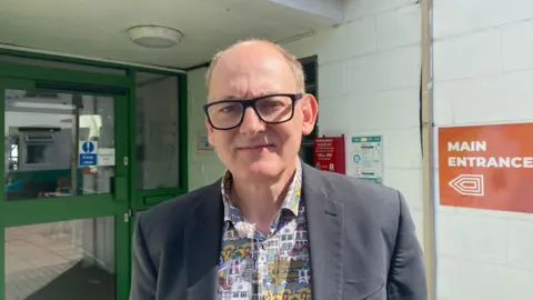 Paul smith wears a grey suit jacket and printed shirt, he is looking at the camera whilst standing outside Easton Community Centre