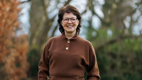 Liam McBurney/PA Wire Shows a woman in a brown jumper, grey trousers with her hands in her pockets. She has dark hair, is wearing glasses and is standing in a garden with trees in the background.