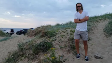 Councillor Dr Tristan Learoyd standing on sand at Marske beach. He has long hair and is wearing sunglasses, a white T-shirt and beige shorts with blue trainers.