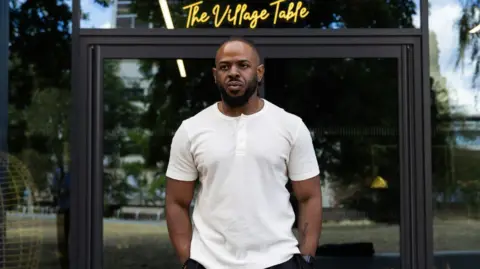 A young black man wearing a white T-shirt and standing outside in front of glass doors