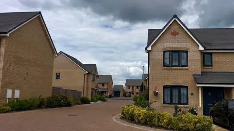 Martin Giles/BBC A clean paved road with a number of new homes on with side. The front house has some yellow and green bushes and a hanging basket.