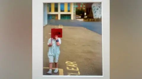 Rachael Carroll A small child wearing a blue and white romper is holding a red flag over their face. Letters on the flag read: "UTB". There are colourful building and a blue and white sculpture in the background, but they are out of focus. The child is walking over yellow road markings.