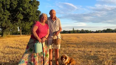 Family handout A couple standing in a field during sunset. They are embracing and smiling at the camera. There is a dog on a lead sitting at their feet. The woman wears a pink T-shirt with a floral white skirt. The man wears a white polo T-shirt and cream shorts. They both hold small buckets. 