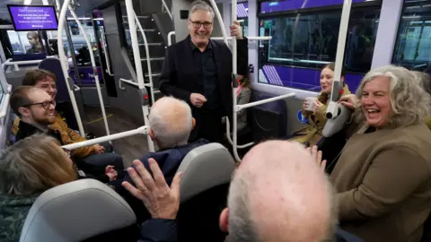 Sir Keir Starmer y Heidi Alexander se ríen con la gente sentada en un autobús. Starmer lleva una chaqueta y camisa oscuras, y Alexander, un abrigo verde.