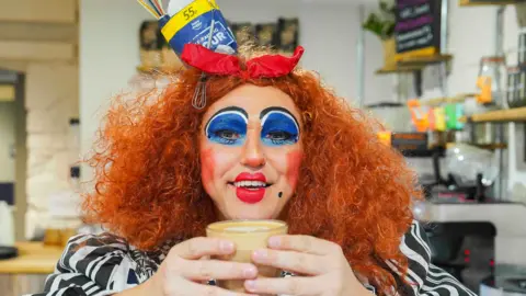 Hugh Hastings A pantomime date with a cup of tea in a glass and wearing a red wig and black and white striped top.