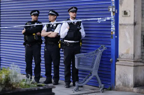 EPA Three police officers behind police tape next to a shopping trolley stand in front of blue shutters