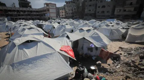 ShelterBox Lots of grey tents to provide shelter to displaced families line a dusty square of land in the middle of damaged buildings. 