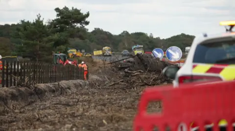 A burnt area of land with piles of cut vegetation. There are works vehicles on a road and a small digger. A person wearing orange high-viz and a white helmet is working on a wooden fence.