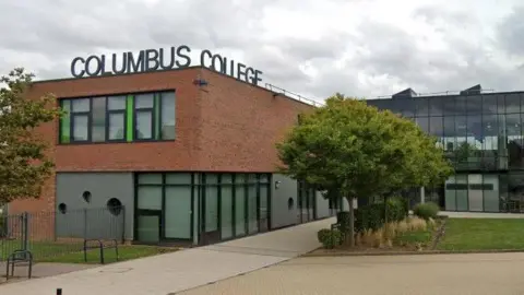 A brown bricked building and glass building is shaped in an L across the image. The bricked building on the left has a sign on the roof which says Columbus College. The glass building on the right has rows or trees outside. 