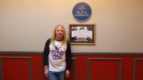 John Quinn Des Rumney with blonde hair, wearing a Black Sabbath long-sleeved top and jeans, stands next to a blue plaque which reads 'Black Sabbath'.