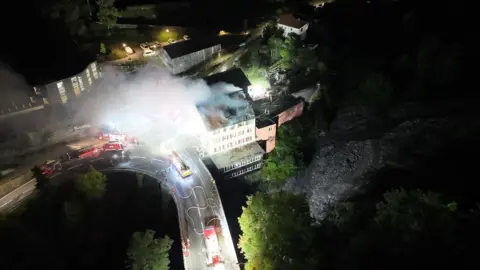 Graubünden cantonal police The building as seen from the air - it is next to a river. Smoke rises from the building at night, while at least five fire engines are parked nearby and on the bridge over the river.