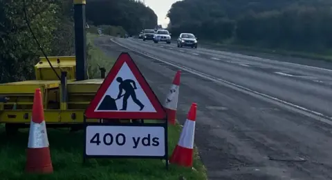 A busy carriageway signs up with red roadworks. There is a white line that indicates a cycle lane.