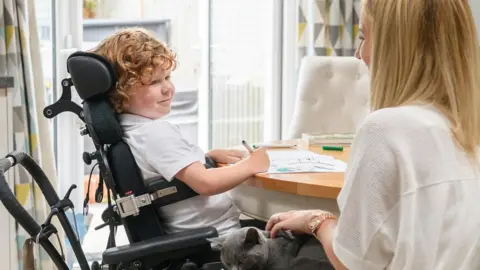 Getty Images A smiling child in a wheelchair sitting at a desk and drawing beside a teacher