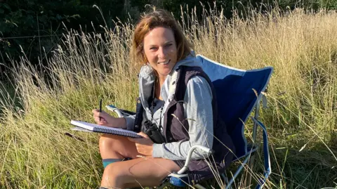 Hannah Dale is sitting in a camping chair in a grassy field, she is holding a notebook and smiling at the cameras. Hanging from her neck is a pair on binoculars, she is also wearing a grey jacket.