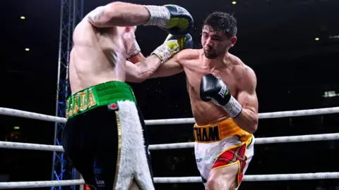 SNS Andy Tham competing in a boxing ring, wearing white shorts with tartan detailing, punching another boxer wearing black and white shorts with green trim.