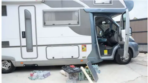 West Yorkshire Police A white and grey motorhome parked on a concrete surface. The motorhome occupies most of the frame, and the passenger-side cab door is open, revealing the driver's area inside, including the steering wheel and front seat. Several items are arranged on the ground beside the open door.