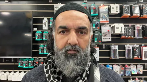 Raz Ahmed, who has a black and grey beard. He is wearing a black-coloured religious headcap.