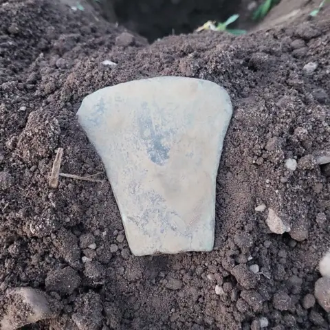 Cameron Anderson Old axe head covered in dust, sitting on dirt, in a field, next to a hole.