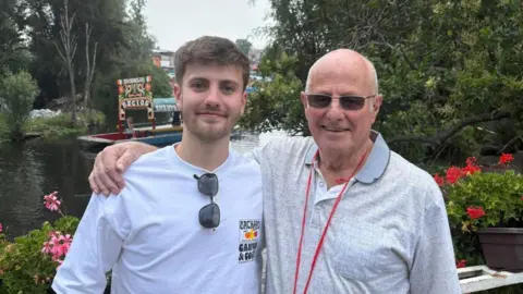 Jacob Allmendinger Two men pose beside a narrow canal lined with trees and flowering plants. One person wears a white long-sleeved shirt with sunglasses, while the other wears a light grey polo shirt, dark glasses, and a red lanyard. A boat is visible on the water in the background.