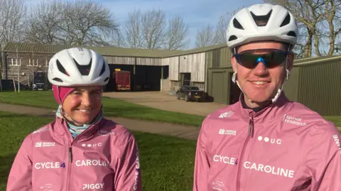 March family Piggy and Tom in cycle helmets - him with sunglasses - with both wearing pink cycling jackets with Cycle 4 Caroline in white lettering across the chest. Other logos in white are dotted around the chest area including Spinal Research and UC