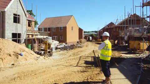A man with a hi-vis yellow jacket and a white hard hat is standing on a building site, looking at new brick homes being built