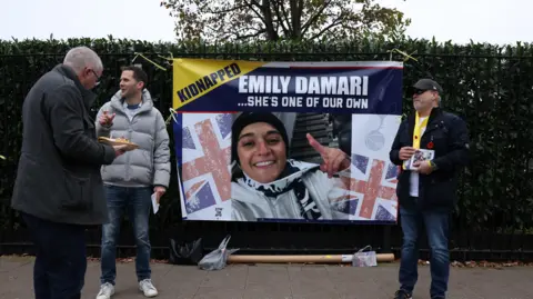 Reuters A large poster with a picture of Emily Damari, union jacks, and the word 'kidnapped' is hung on a hedge. Three men stand beside it, one with pamphlets in his hand.