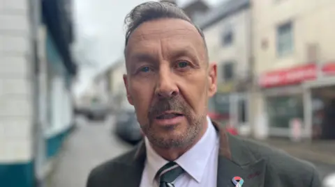 The head and shoulders of a man with greyish-brown hair and beard looking directly at the camera, wearing a grey suit with a brown collar, a striped tie and an LGBT+ veterans pin badge