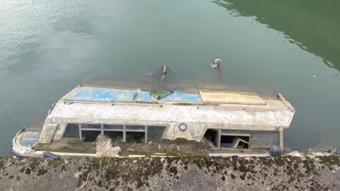 A boat partially submerged in the river in Bath. It is a large boat with debris to it's exterior and smashed windows.