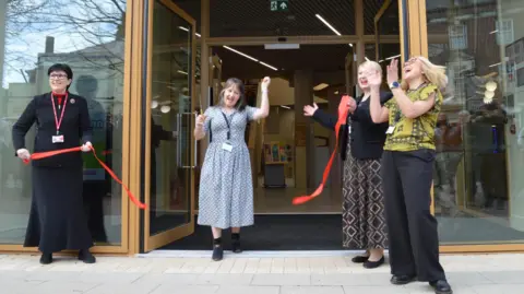 Norfolk County Council Four people are standing in front of the library at the entrance. A person is standing in the middle and has just cut a red bit of ribbon. Two people on the right are clapping and a person is on the left holding the red ribbon.