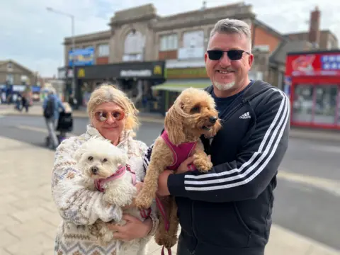 Owen Sennitt/BBC A man and a woman smiling and looking towards the camera while holding two small dogs in their arms.