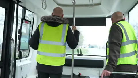 Two men inside a moving self-driving bus. They are both wearing high visibility jackets and facing away from the camera.