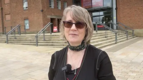 A photo of a woman looking into the camera she has got short brown hair and is wearing a small fabric scarf around her neck
