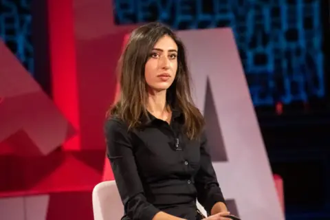 Getty Images A young woman with long brown hair, dressed in a black shirt, sits on a white chair in a TV studio