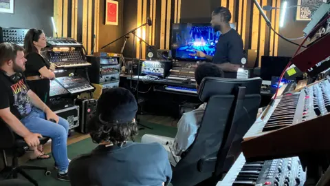 A man in a recording studio stands in front of a computer. There are four other people, sitting down watching him. There are mics and keyboards.