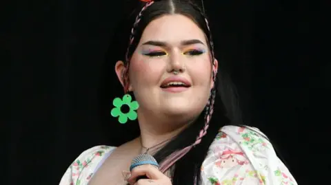 Getty Images Mae Stephens has multi-coloured eye shadow and is wearing large green flower earrings. She has a pink plait in her long brown hair and is holding a microphone. The background is black.