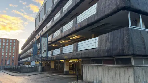 Exterior of a concrete multi-storey car park, five floors are visible. A hanging sign says "Welcome to Chapel Street Cathedral Quarter"
