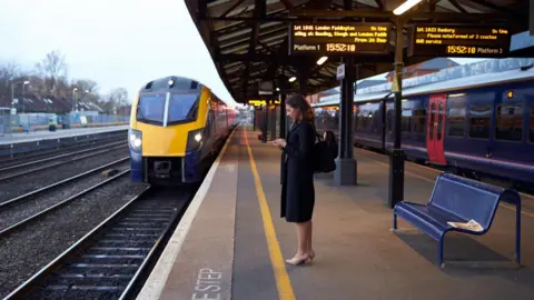 A woman is on her phone while she waits on a train platform. There is a yellow and blue train on the tracks in front of her