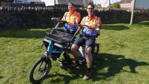 John and Edouard sitting in a field on a side-by-side tandem wearing hi-vis orange jackets and shorts, smiling to camera