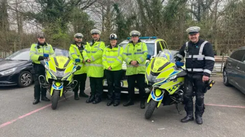 Several officers from the Tasking Team posing for a picture. A police car and two police motorcycles are beside them. It is an overcast day.