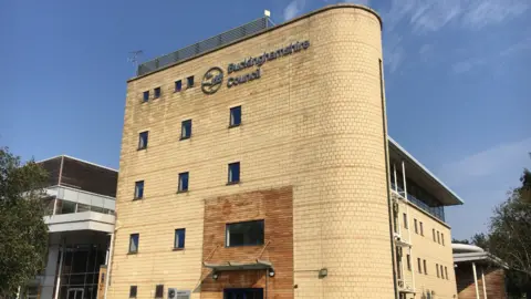 A picture of Buckinghamshire Council's gateway building in Aylesbury. It is a large yellow brick building, which is five stories high. It has a hedge in front of it, and the sky is blue behind it.