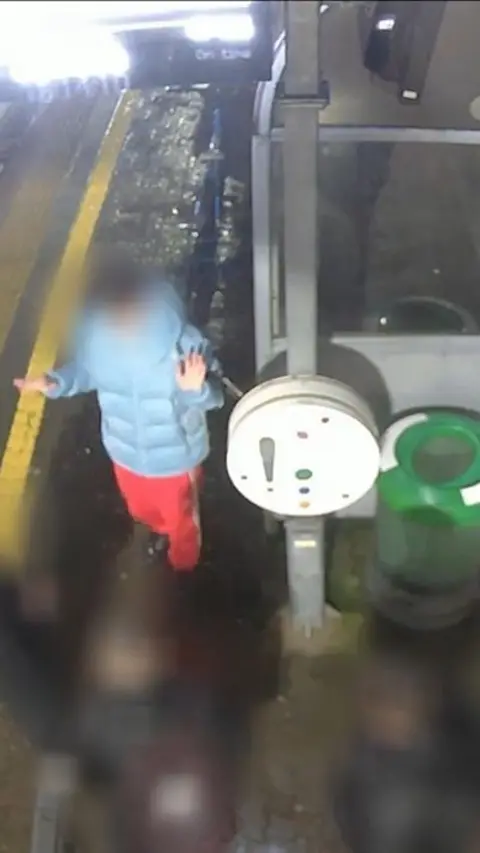 A boy with a blurred face in a blue coat and red trousers on a railway platform.