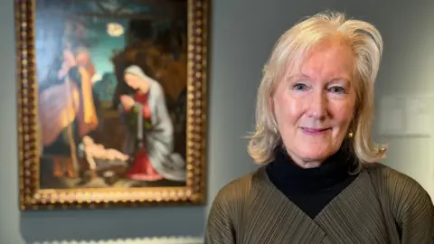 A woman with blond hair is standing in front of a painting of the nativity scene. The woman is wearing a black jumper with a green cardigan on top. 