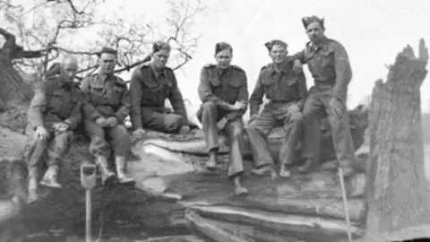 British Resistance Museum (from left to right) Sgt Arthur Dickerson, unknown man, either Roy or Gerald Allen, Geoffrey Newman,  either Roy or Gerald Allen (they were brothers) and  Cpl Herbert Lynes. The photo is a blurry black and white and shows a row of men, wearing Home Guard uniforms, and sitting on a huge tree trunk.