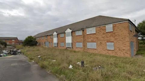 An exterior view of Admiral Court Nursing home. The building is a long brick structure with windows with white window frames. 