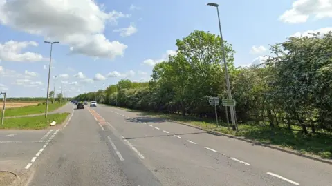 A Google Streetview shot of the A428 on a sunny day. There are fields on the left and a line of trees running alongside the road on the right.