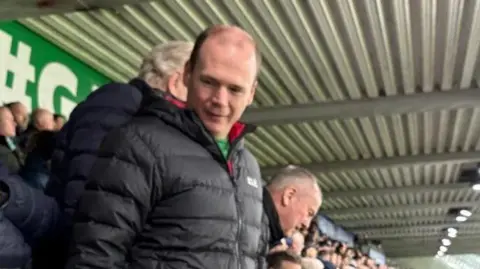 BBC Minister Lyons standing in the stands at the match wearing a black puffer coat.