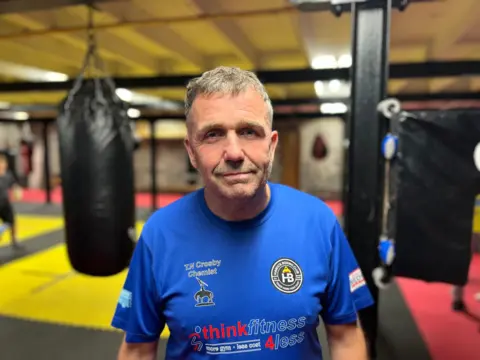 Un hombre de cabello gris, con una camiseta azul, mira la cámara. Detrás de él hay un gimnasio de boxeo
