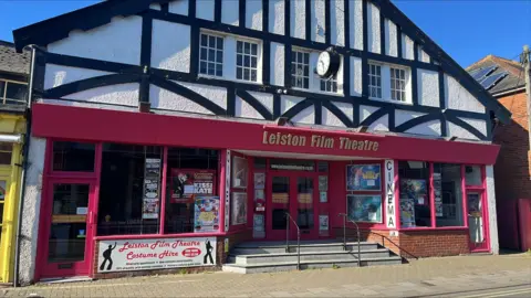 A general view of the exterior of the Leiston Film Theatre. The upper half of the building has been built in a Tudor style. The bottom half has red beams and outlines along with the name of the theatre above its entrance.